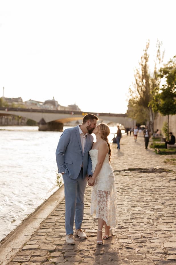 Bride and groom kiss on a cobblestone Paris riverbank in warm evening light, with a bridge behind them