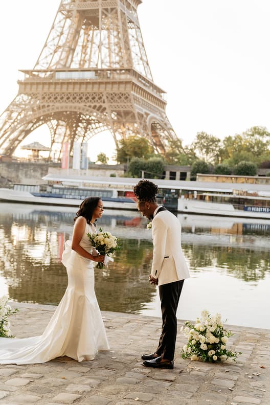 Bride and groom share vows by the Eiffel Tower in warm light, with white bouquets and calm water behind them