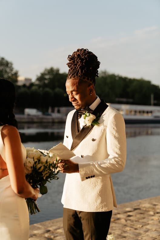Groom in a cream tux reads vows as the bride holds a bouquet on a Paris riverbank at sunset