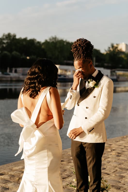Bride and groom stand by the river on a Paris street, the groom dabbing tears in warm evening light