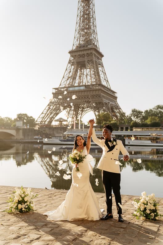 Bride and groom raise joined hands by the Eiffel Tower beside the Seine, with petals in sunlight