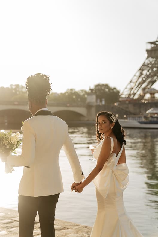 Bride and groom hold hands by the Seine at sunset, with the Eiffel Tower behind them