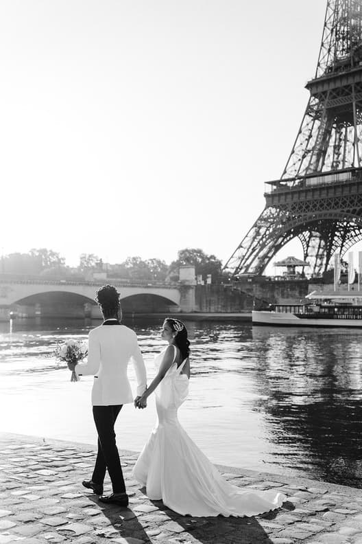Bride and groom hold hands by the Seine in bright backlight, with the Eiffel Tower behind them