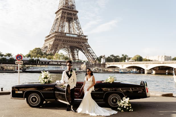 Bride and groom pose beside a black vintage car by the Seine, with the Eiffel Tower behind them in soft daylight