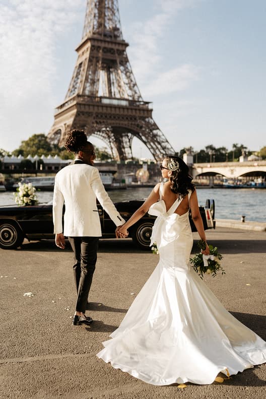 Bride and groom walk hand in hand by the Seine with the Eiffel Tower behind them in warm light