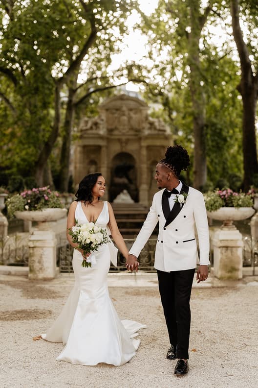 Bride and groom hold hands in a shaded garden, with a stone fountain and soft afternoon light