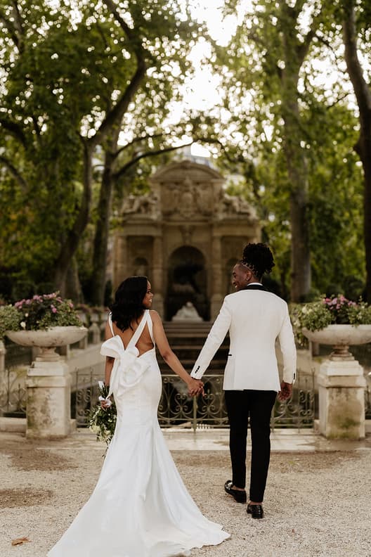 Bride and groom in white hold hands in a tree-lined garden, backlit by soft daylight and framed by stone urns