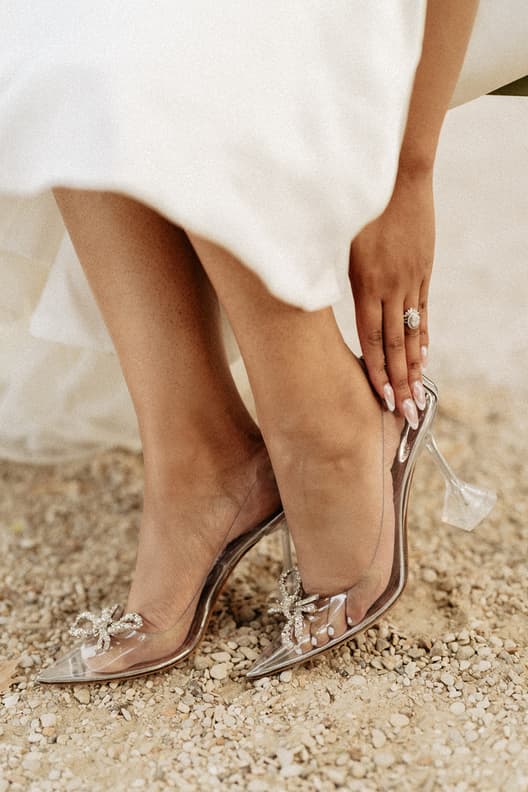 Bride in clear heels with jeweled bows touches her hem on a gravel path in warm light