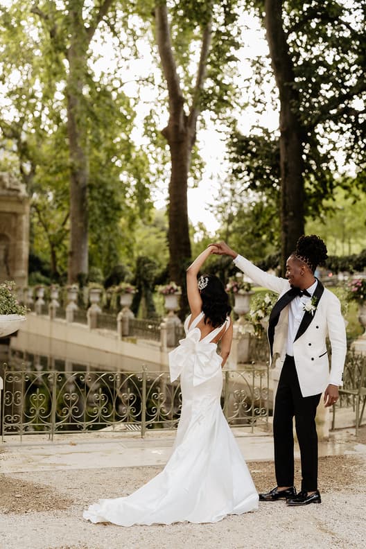 Bride and groom dance in a sunlit garden, her gown’s bow trailing behind her