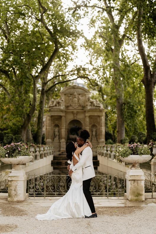 Bride and groom embrace in a tree-lined garden before a stone fountain and wrought-iron railing
