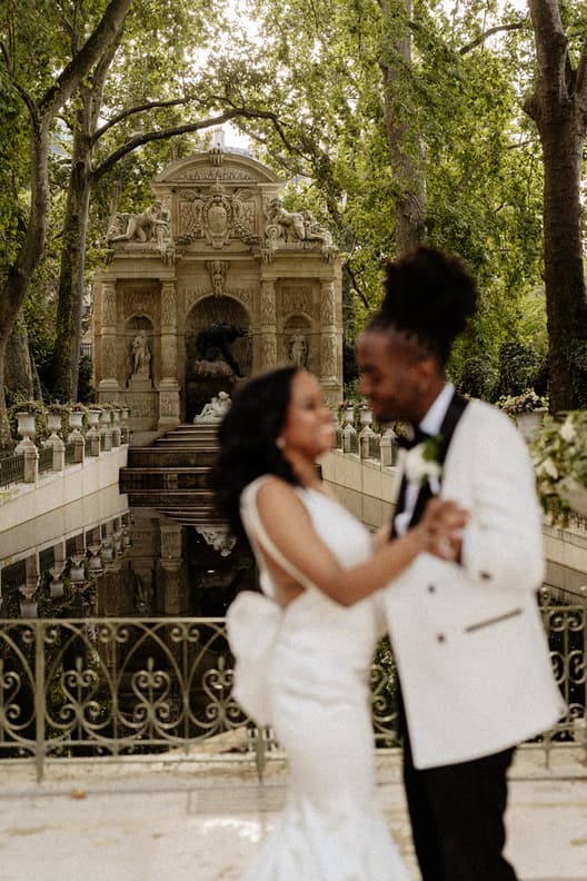 Bride and groom dance in a garden before an ornate stone fountain and reflecting pool under soft daylight