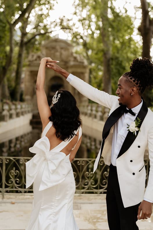 Bride and groom twirl on a tree-lined bridge in soft light, with a bow-backed gown and tuxedo