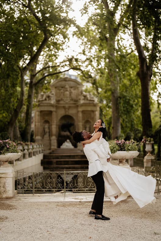 A groom lifts his bride in a sunlit Paris garden, with stone steps and pink flowers behind them