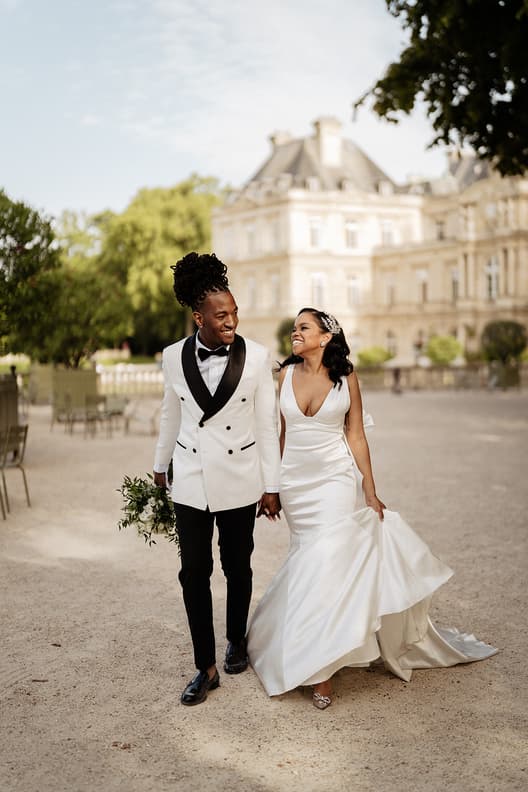 Bride and groom walk hand in hand through a Paris garden in soft light, smiling by a chateau façade