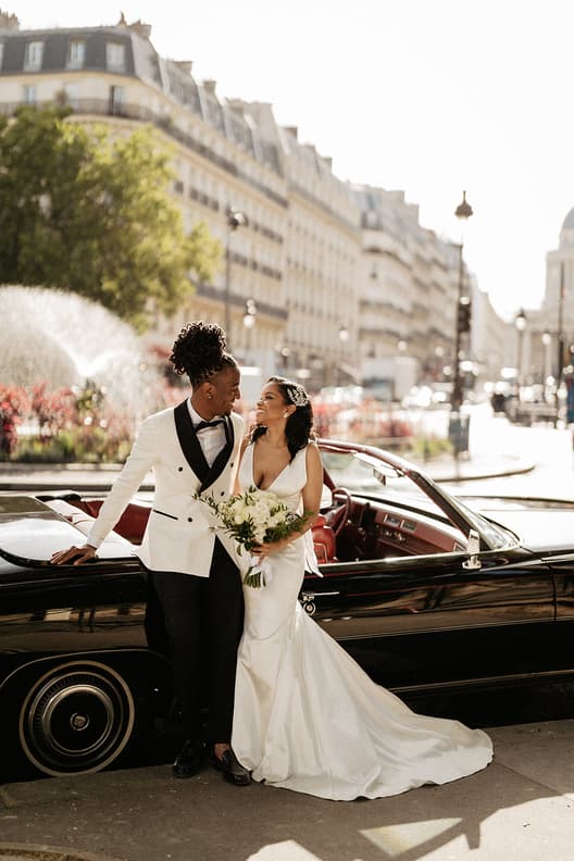 A bride and groom pose beside a black convertible on a bright Paris street, sharing a bouquet