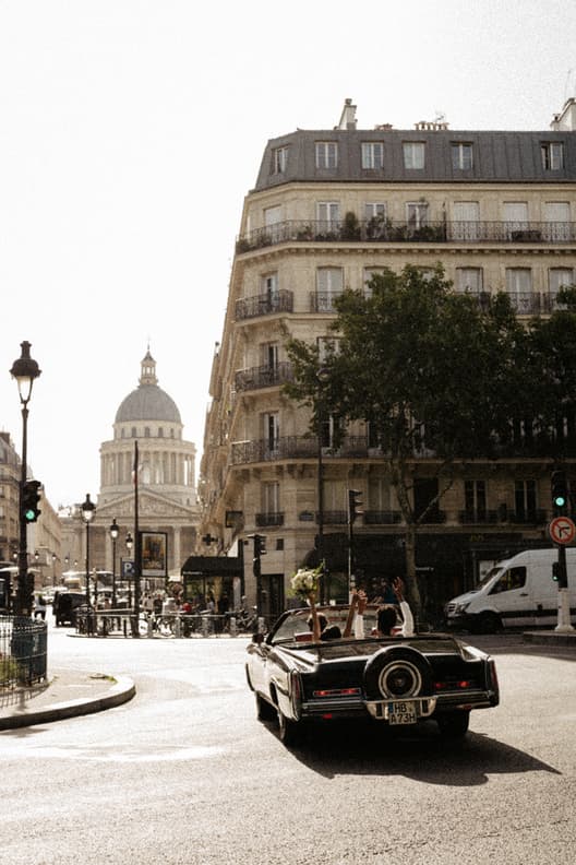 A couple rides through a Paris street in a black convertible, arms raised in warm afternoon light