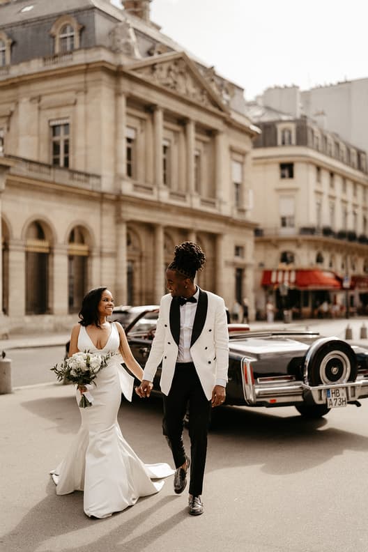Bride and groom hold hands on a Paris street beside a classic car in warm daylight