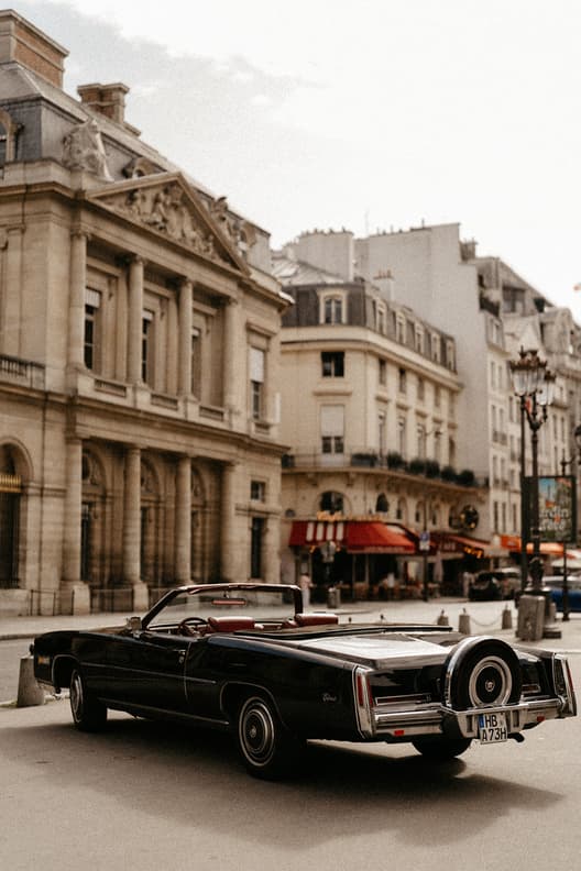 A black vintage convertible rests on a Paris street beside Haussmann buildings in soft daylight