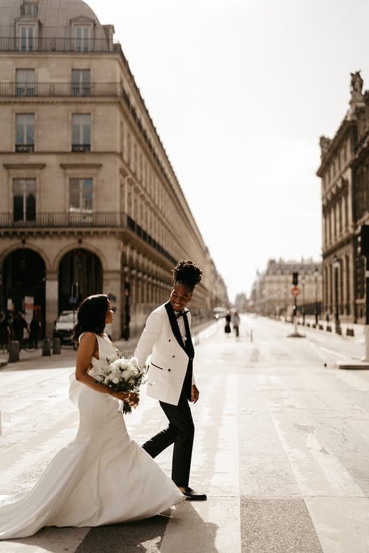 Bride and groom walk hand in hand on a Paris street in warm light, her bouquet and long train flowing