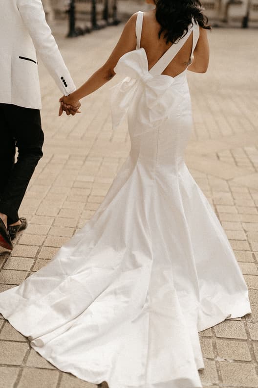 A couple in wedding attire holds hands on a Paris street, with a long white train trailing behind in soft daylight