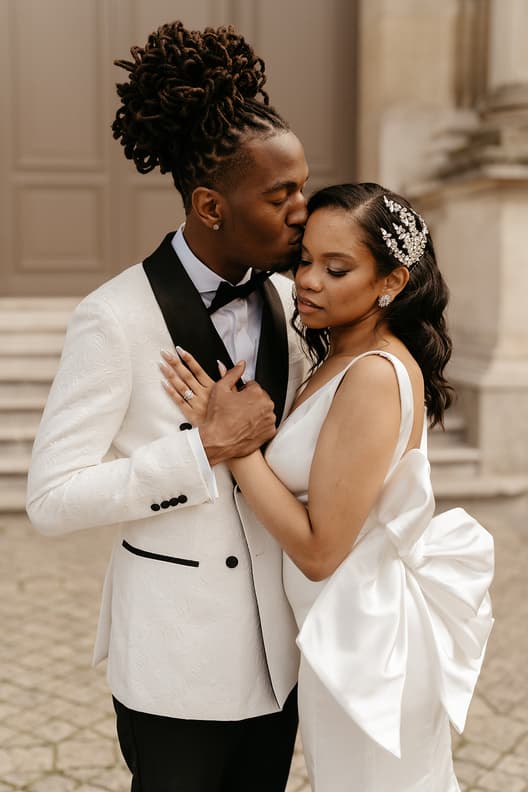 Couple in wedding attire share a tender forehead kiss beside a stone Paris street