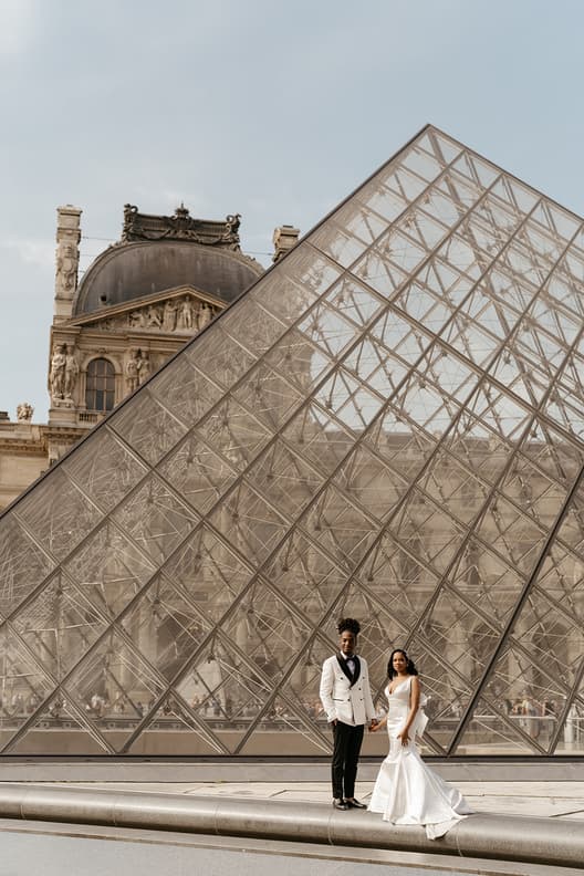 Bride and groom in wedding attire stand before the Louvre pyramid in soft daylight