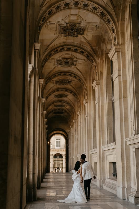 Bride and groom walk hand in hand through a vaulted Paris arcade in soft golden light