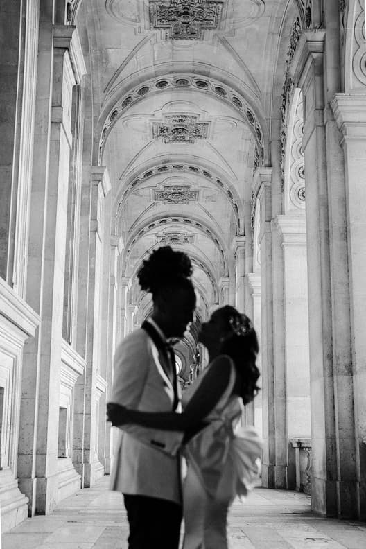 Couple embraces in a grand stone arcade, soft daylight tracing the arched ceiling and her bow gown