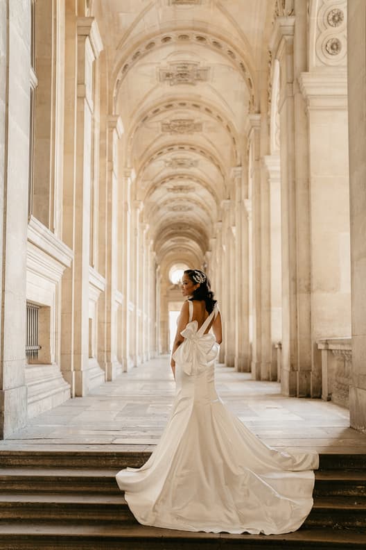 Bride in a flowing gown turns in a sunlit Paris colonnade with a long train