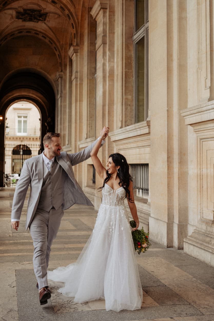 Bride and groom in formalwear walk arm in arm through a Paris arcade, with warm light and a bouquet in hand