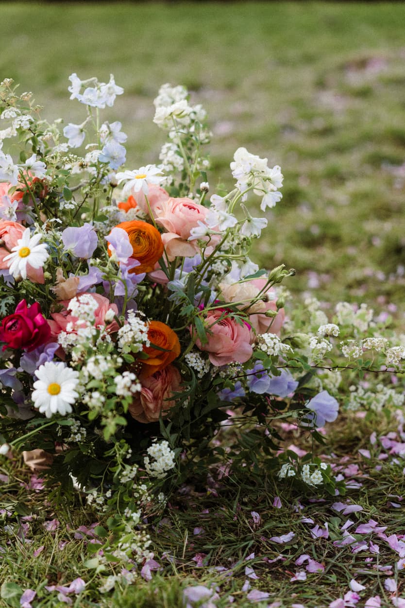 A colorful bridal bouquet of roses and daisies rests on grass scattered with petals