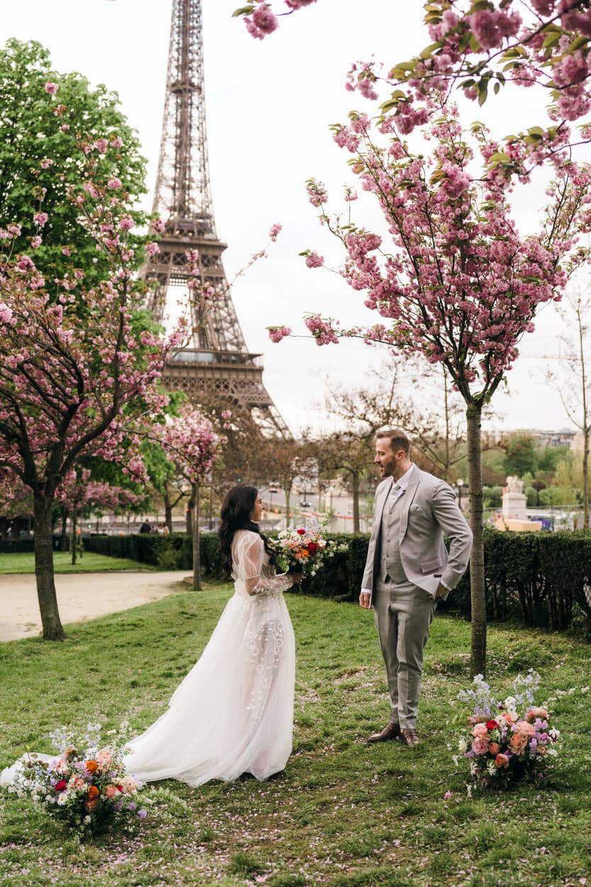 Bride and groom stand in a flowered garden by the Eiffel Tower in soft daylight