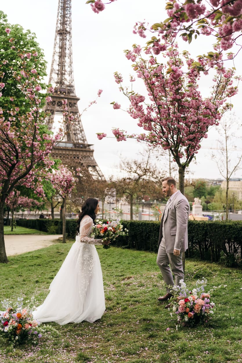 Bride and groom face each other in a blossom-filled garden with the Eiffel Tower behind them