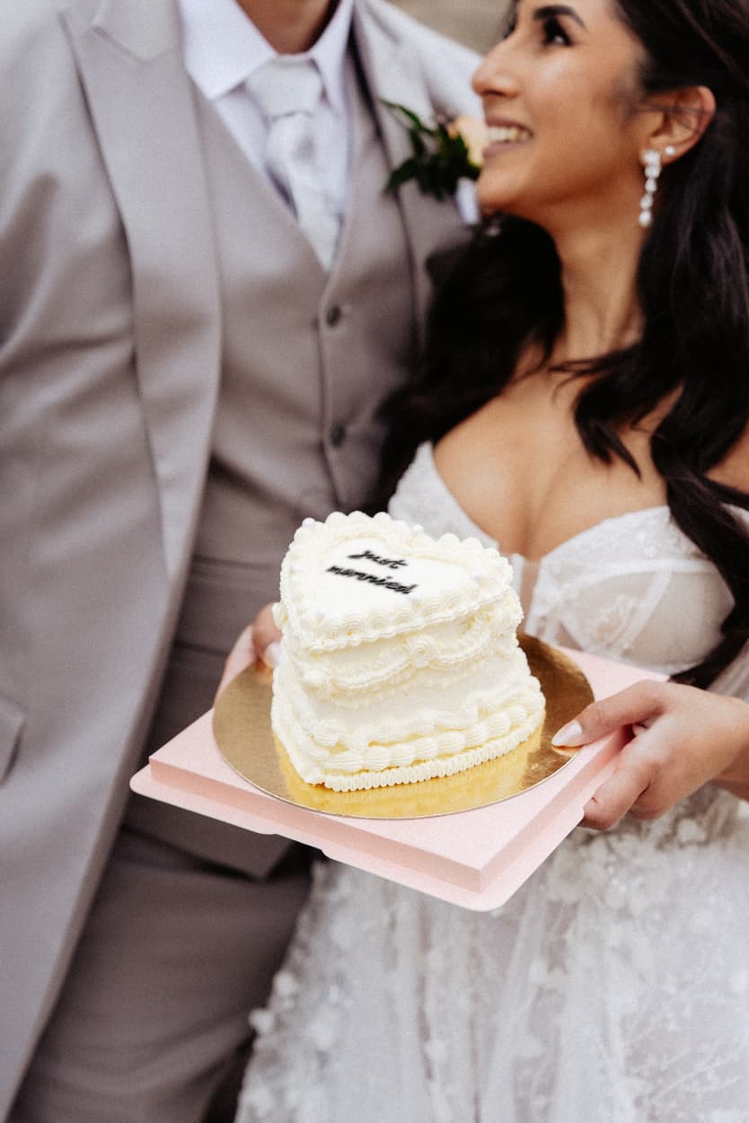 Bride and groom hold a small white cake on a pink tray in soft light, close together
