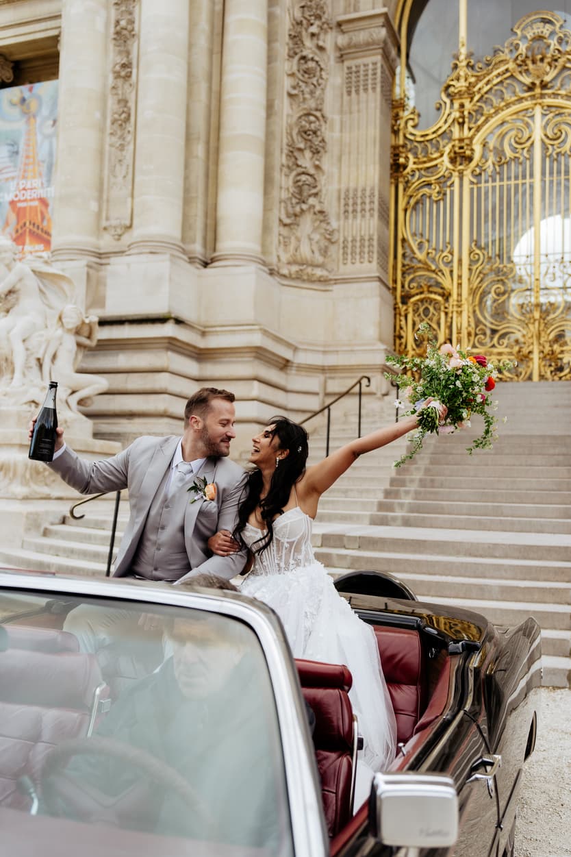 Bride and groom cheer in a vintage car before an ornate Paris building in soft daylight