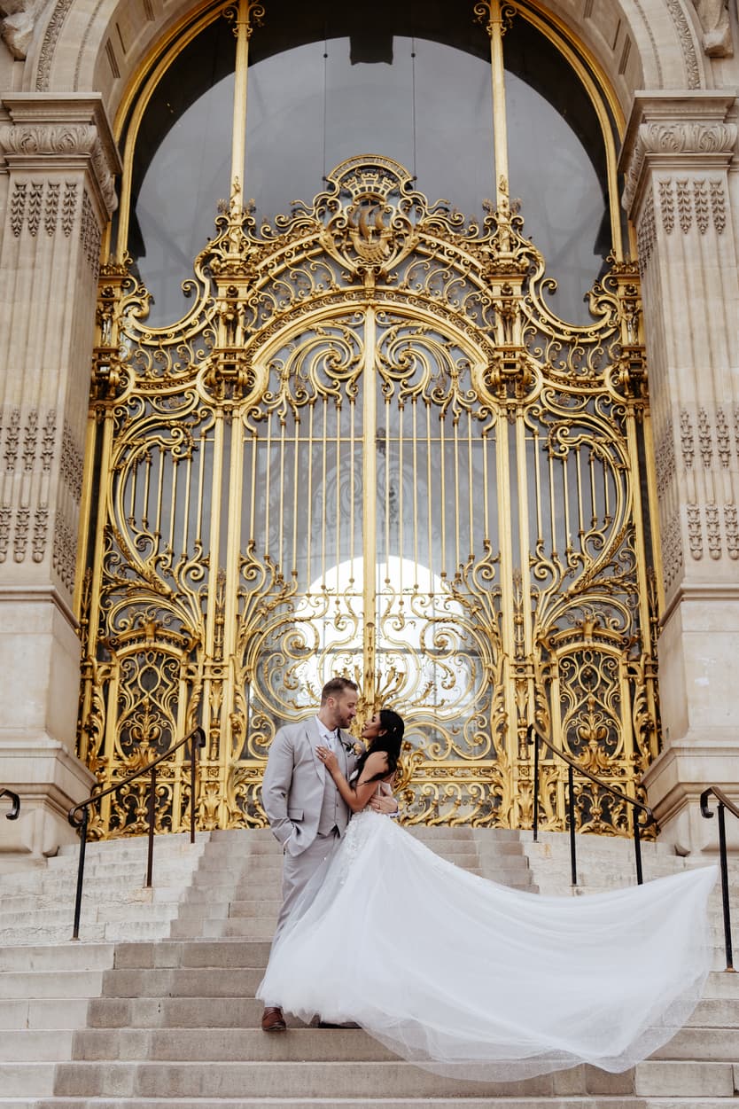 A bride and groom embrace on stone steps before an ornate gilded Paris entrance