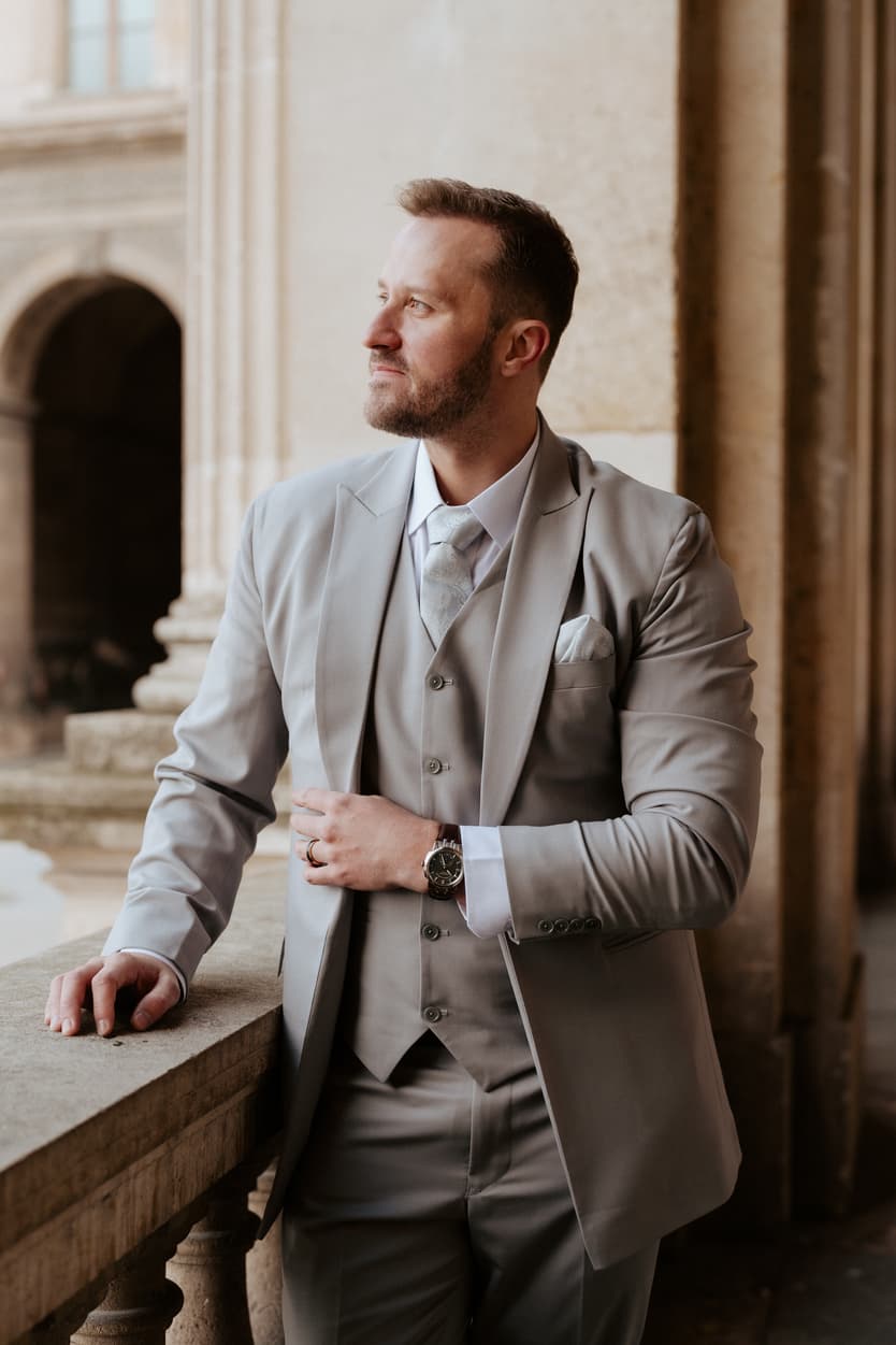 Groom in a light gray suit leans on a stone balcony in warm light beside a Paris arcade