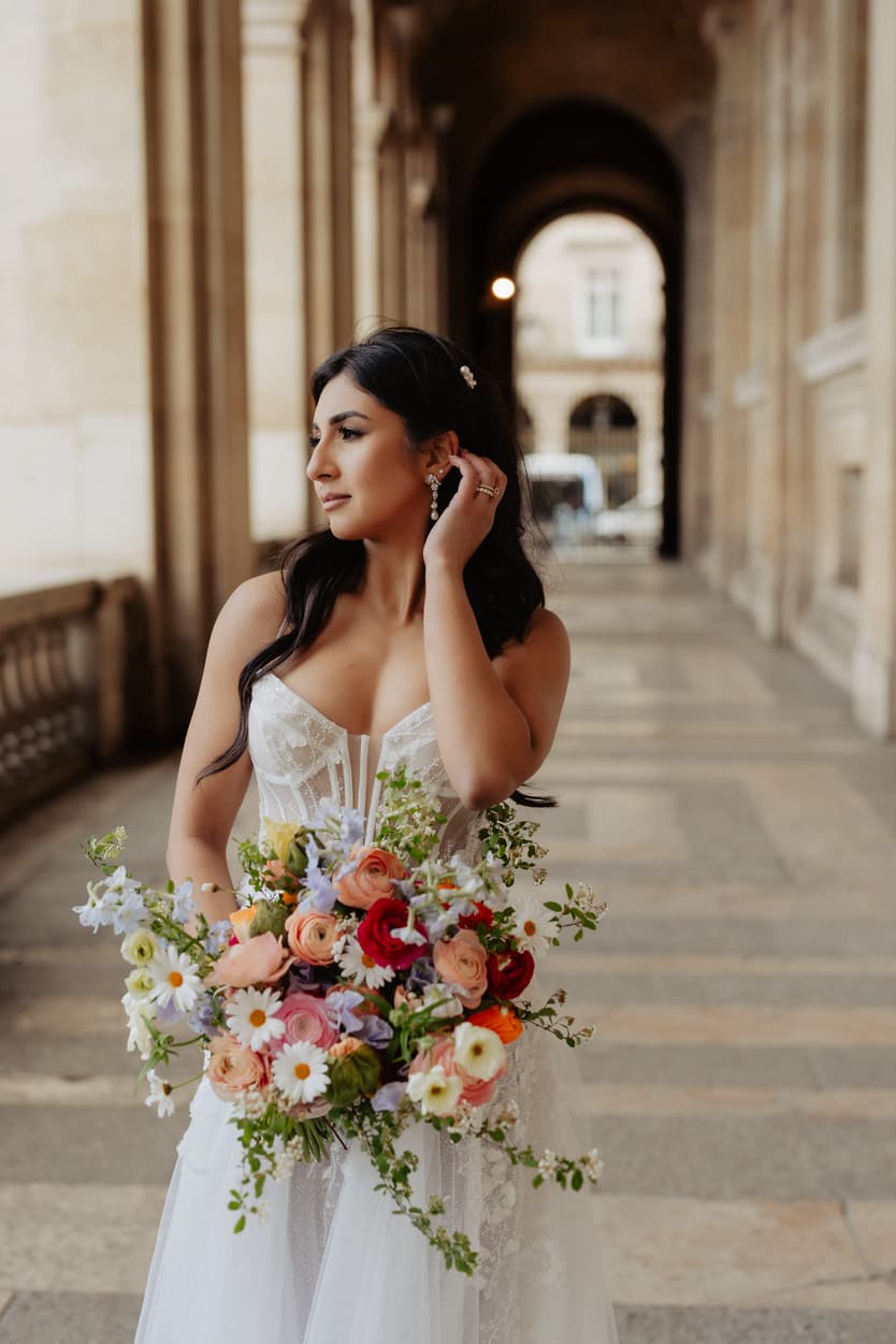 Bride in a white dress holds a colorful bouquet beneath a Paris stone arcade