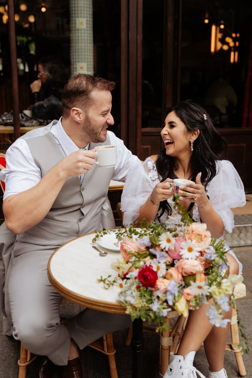 Bride and groom laugh over coffee at a Paris cafe, with a bright bouquet on the table