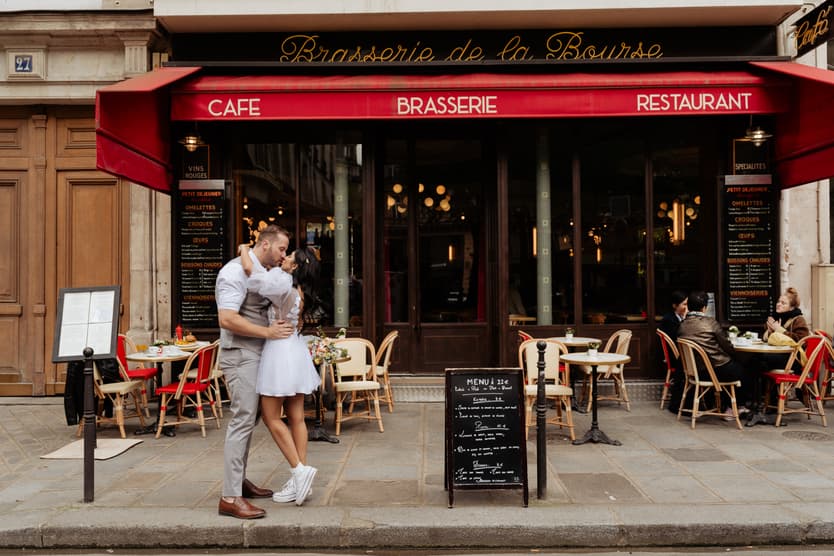 A couple embraces outside a Paris brasserie with red awnings, café tables and warm window lights