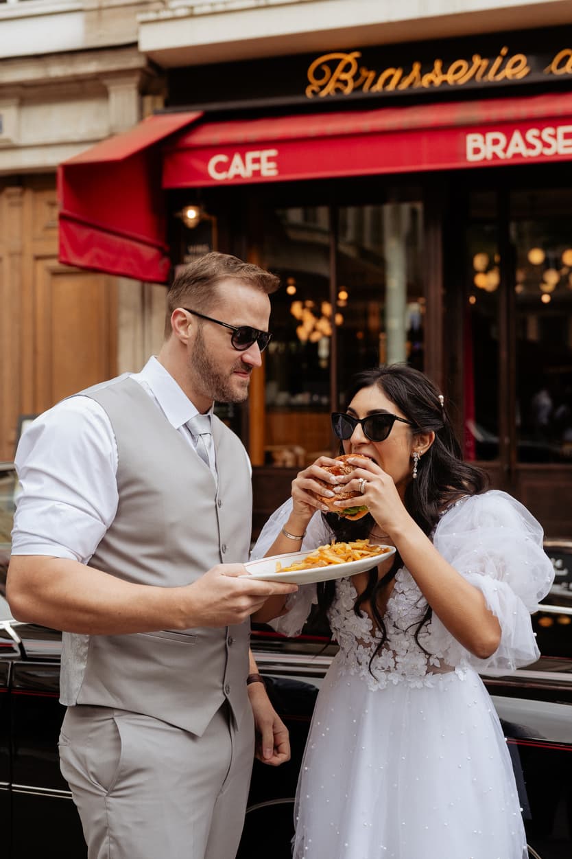 A bride and groom share a burger and fries outside a Paris street café under a red awning