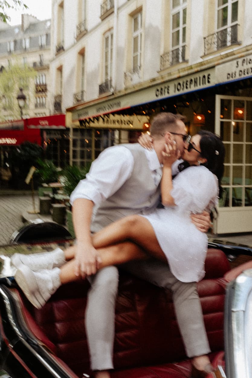 A couple kisses in a vintage car on a Paris street outside Café Joséphine, with warm cafe lights glowing