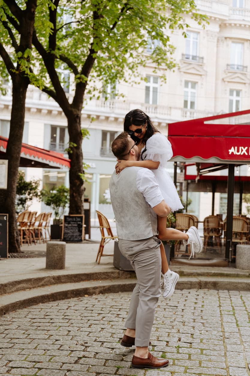Couple embraces in a Paris street café, the groom lifting the bride under leafy trees and red awnings