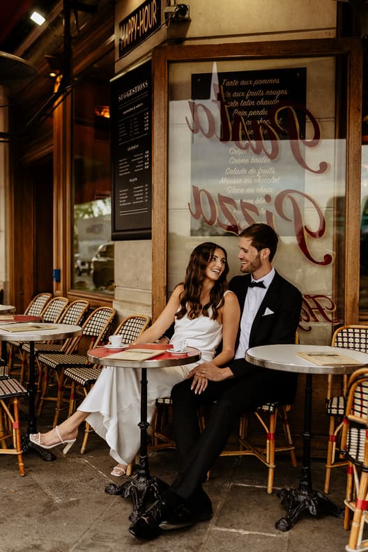 Bride and groom sit together at a Paris street cafe, smiling in warm light beside woven chairs