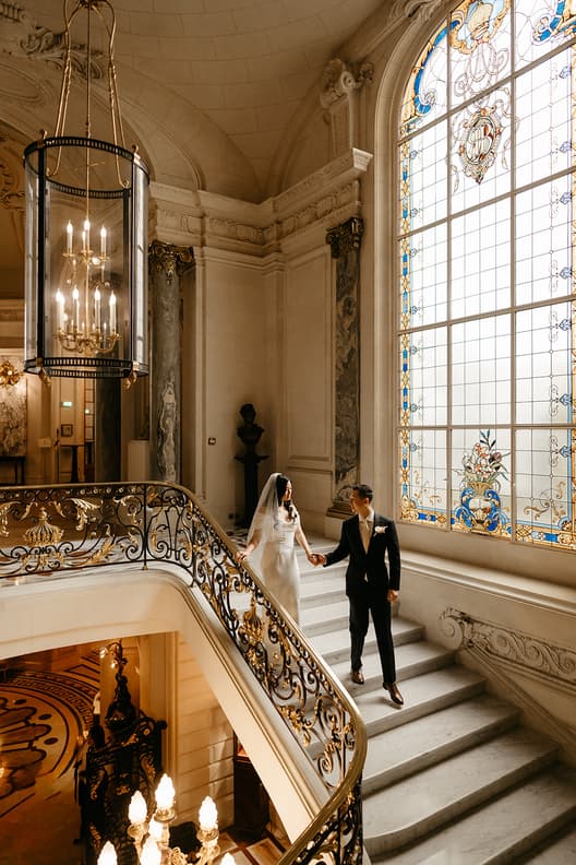 Bride and groom hold hands on an ornate staircase beside a stained-glass window in warm light