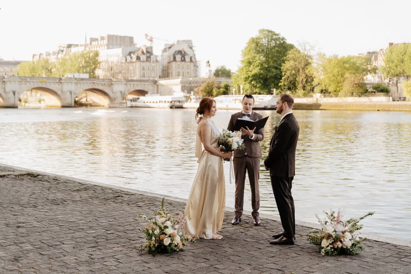 Bride and groom stand with an officiant by the Seine, framed by bouquets and soft evening light