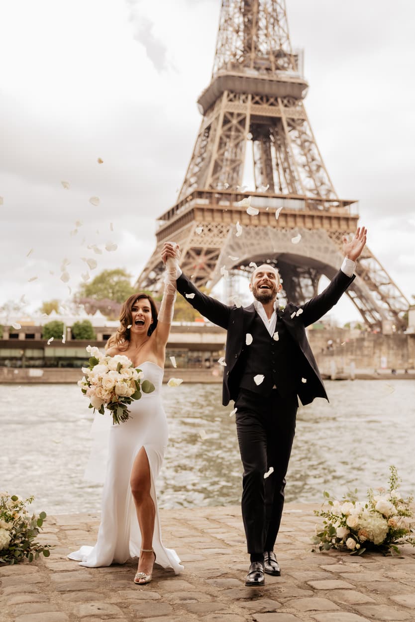 Bride and groom celebrate with raised arms and confetti by the Eiffel Tower on a cloudy Paris riverside