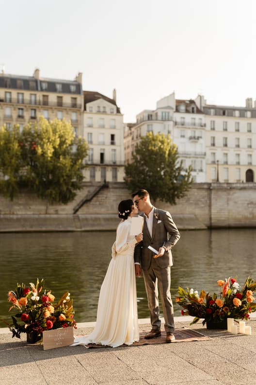 Bride and groom kiss by the Seine beside floral arrangements in warm light