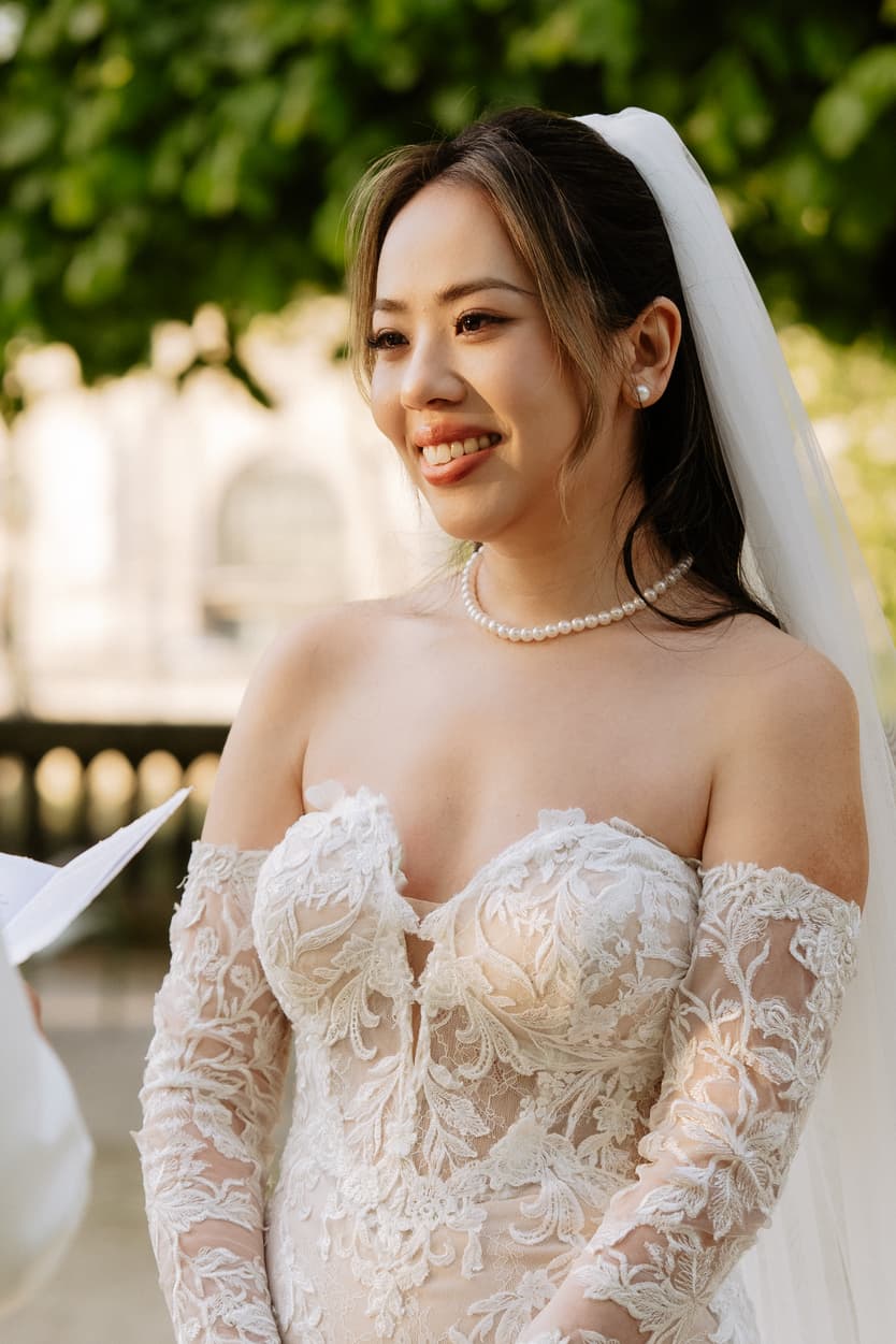 Bride in lace gown and veil smiles in soft light beside a stone balustrade in a garden