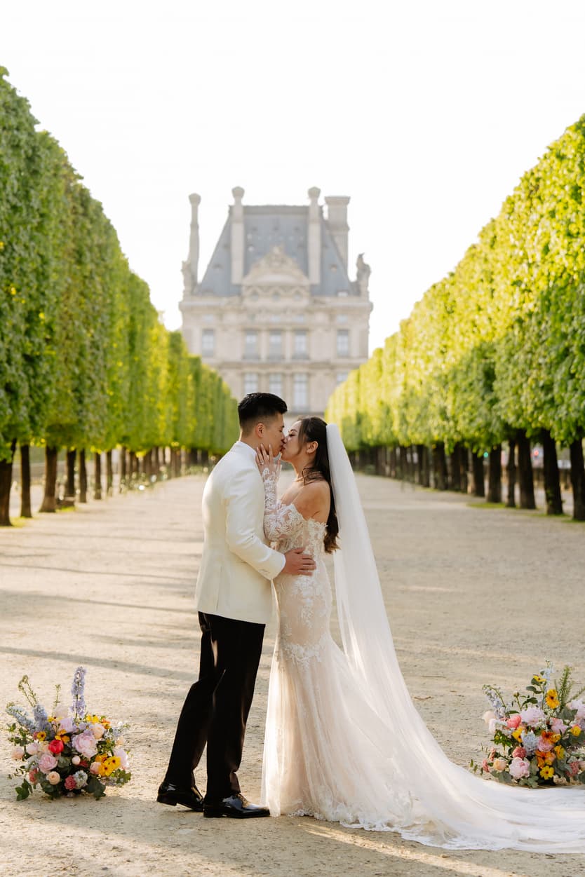 Bride and groom kiss in a Paris garden, framed by clipped trees and soft afternoon light